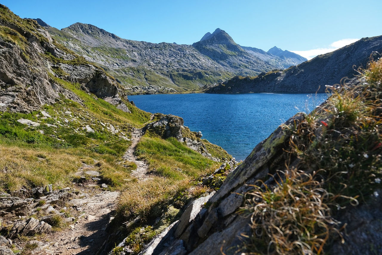 Wanderweg am Ufer eines Bergsees mit tiefblauem Wasser und Felsgipfeln im Hintergrund