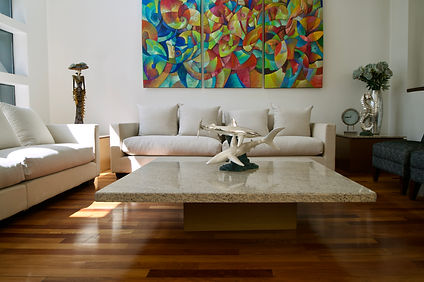 This image shows a contemporary living room with a rich wooden floor and a large, colorful abstract painting dominating the wall above a minimalist white sofa. In the center, a coffee table displays a unique sculpture, contributing to the room's modern and artistic atmosphere.