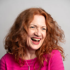 Joanne has long, wavy red hair and is wearing a bright pink jumper with silver pendant. She sits directly on to the camera, laughing, and looking out to the right of the frame.