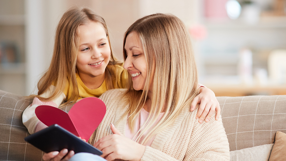 Mother and daughter sitting on a sofa reading together, child holding a pink heart for Valentine’s Day, cosy family learning moment at home.