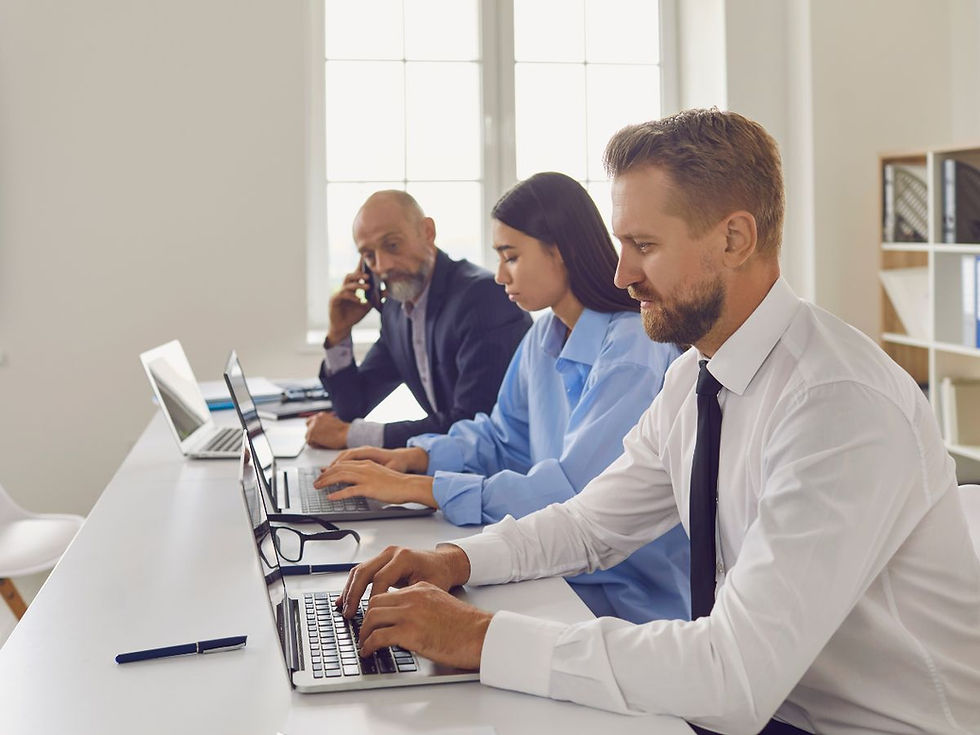 Man in a suit looks pensive near a window. In the blurred background, four people hold mugs, smiling, suggesting a workplace setting.