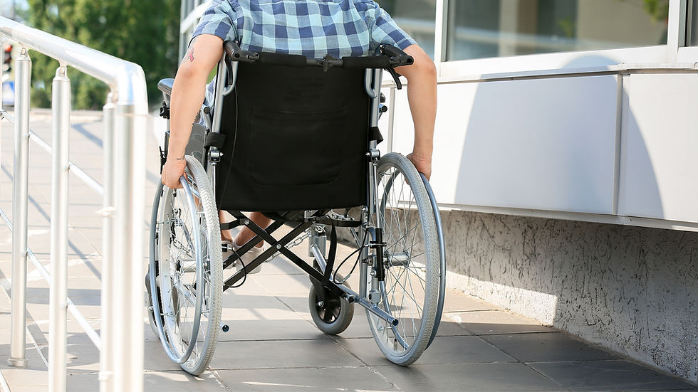Person in wheelchair, wearing a plaid shirt, moves up a ramp outdoors. Metal railing visible, sunny day, concrete building in background.