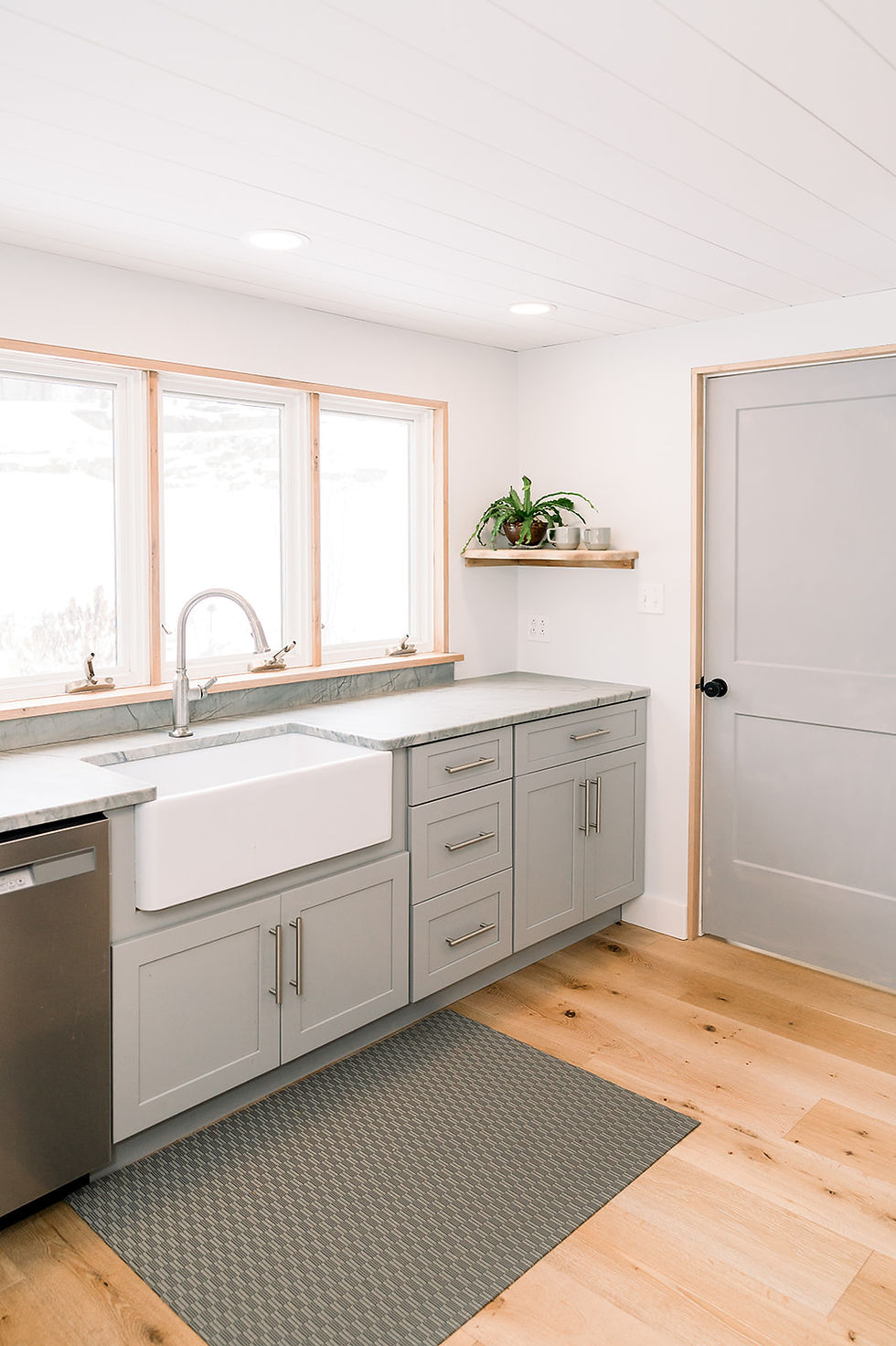 A stunning modern farmhouse kitchen featuring soft gray cabinetry, a large apron-front sink, natural wood trim, and open shelving adorned with plants. Light floods in through large windows, enhancing the airy aesthetic.