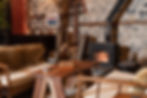 A detailed view of a black wood stove with a fire burning inside, set against a stone wall. A live-edge wooden coffee table sits in front of the fire, surrounded by wooden-framed chairs with cream cushions.