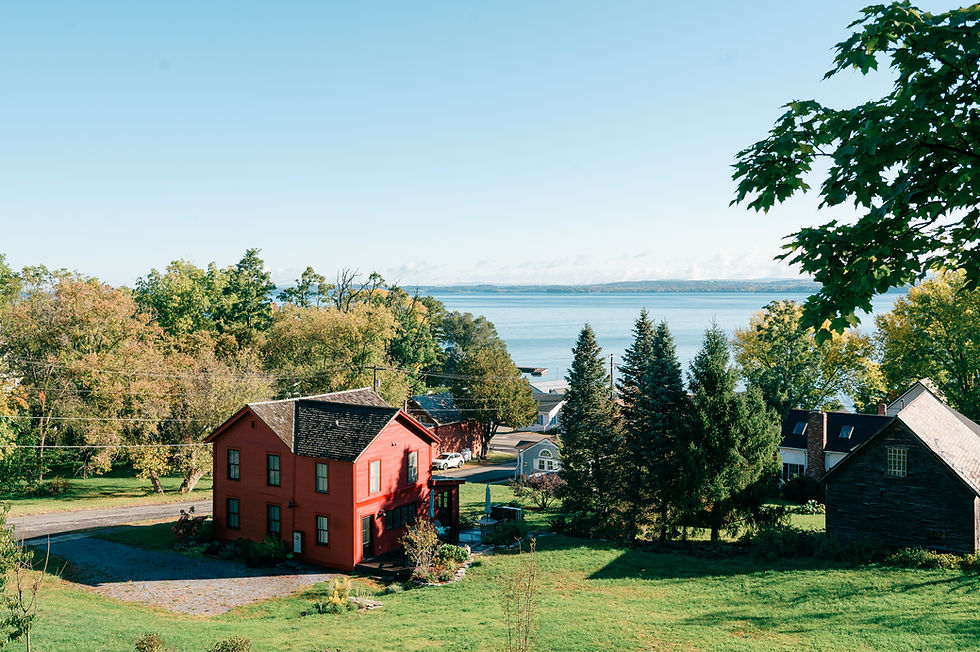 A scenic view of a restored historic home with a red facade and dark roof, nestled among lush greenery overlooking Lake Champlain. The property features a well-maintained lawn, additional buildings, and a backdrop of trees with autumn foliage. Sawteeth Carpentry specializes in historic restorations, custom remodels, and new home builds in upstate New York, Essex, and the Lake Champlain region.