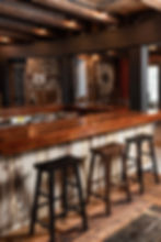 A handcrafted bar made of wood and reclaimed materials, featuring black-painted support beams. Four rustic wooden stools sit along the bar, and the background showcases exposed brick walls, a dartboard, and wooden tables.