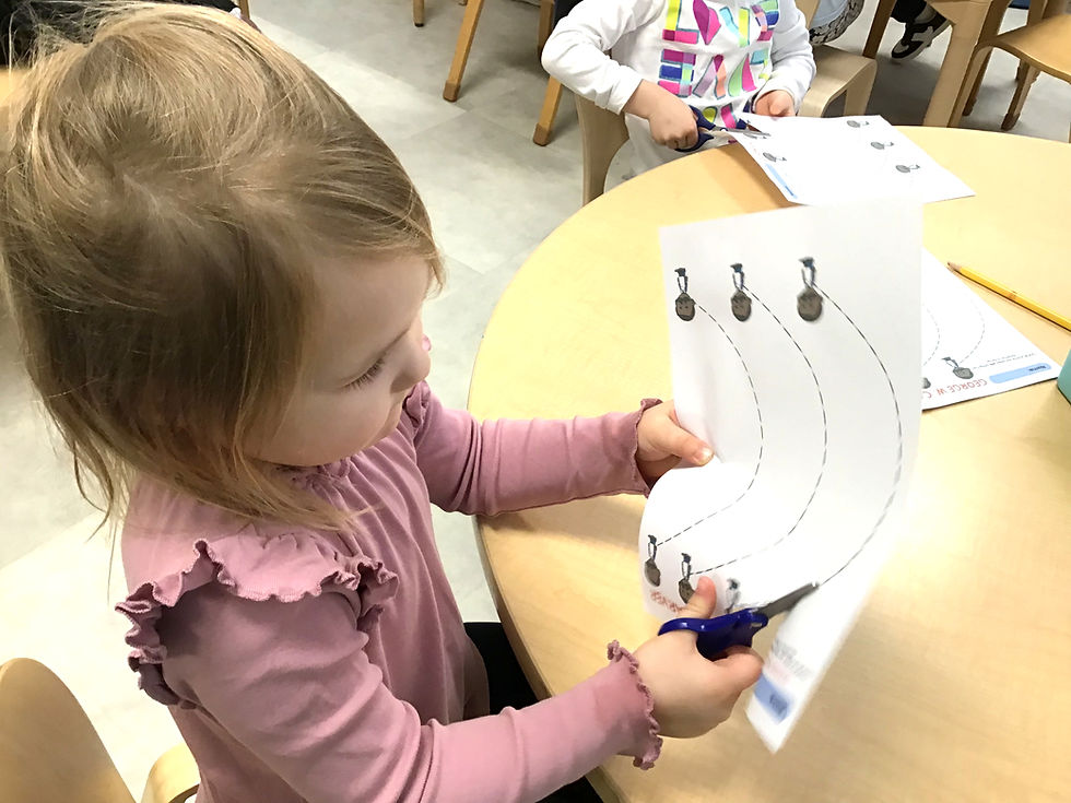 Child in pink shirt uses scissors to cut along curved lines on paper at a table. Another child in colorful shirt also cuts paper nearby.