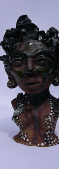 Sculpture of a woman wearing sizeable black pearl earrings. Her skin is made of dark clay—with a green glaze on the top half of the bust. She wears decorative paint on her body. She has short curly hair. Made by Luisa Manea