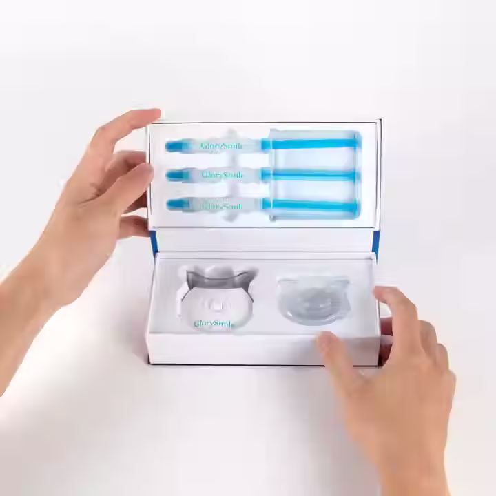 A person holding an open teeth whitening kit labeled "GlorySmile." The kit includes three blue gel syringes in the top compartment and a white LED light device with a clear mouth tray in the bottom compartment