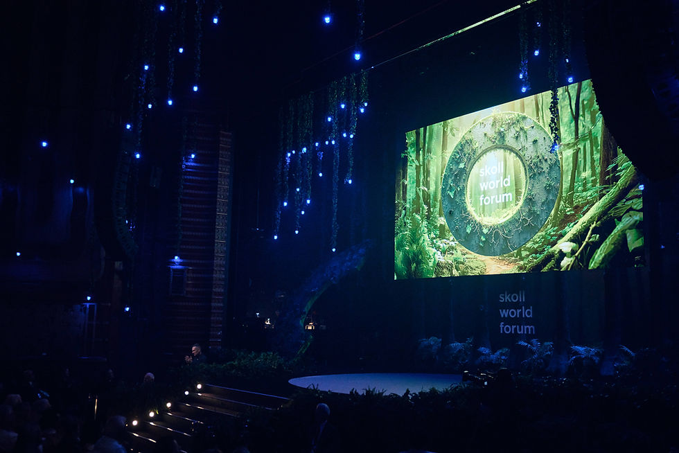 Dimly lit stage with blue lights and green foliage. Large screen displays "Skoll World Forum" in forest setting. Audience visible.