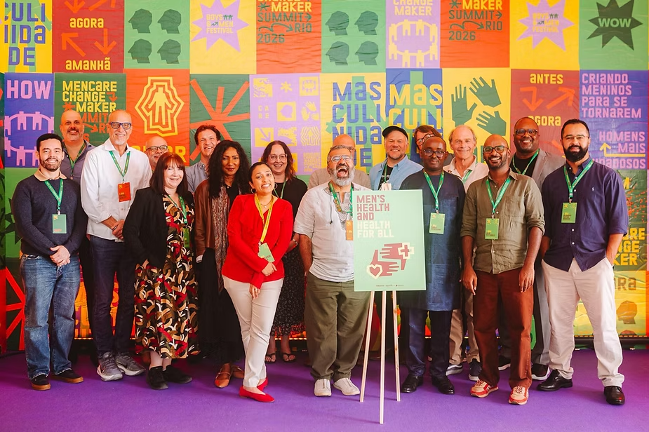 The RWAMREC ED and other change makers in the men's health track posing on a purple floor before a colorful festival wall, holding a sign reading Men’s Health and Health for All.