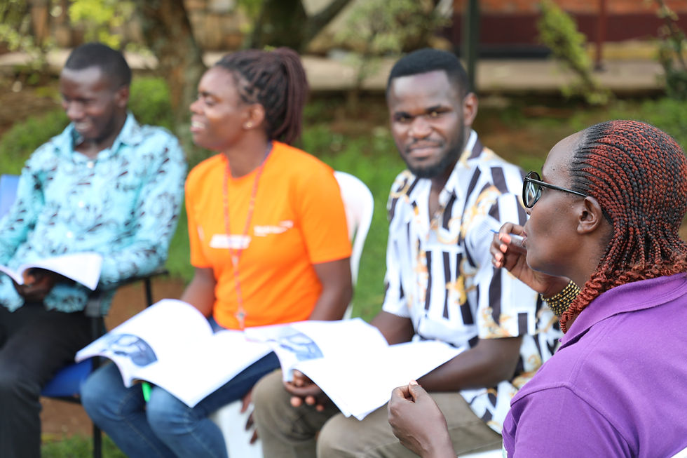Four people sitting outdoors, holding RWAMREC modules, engaged in discussion. One wears a bright orange shirt. The background shows greenery and trees.