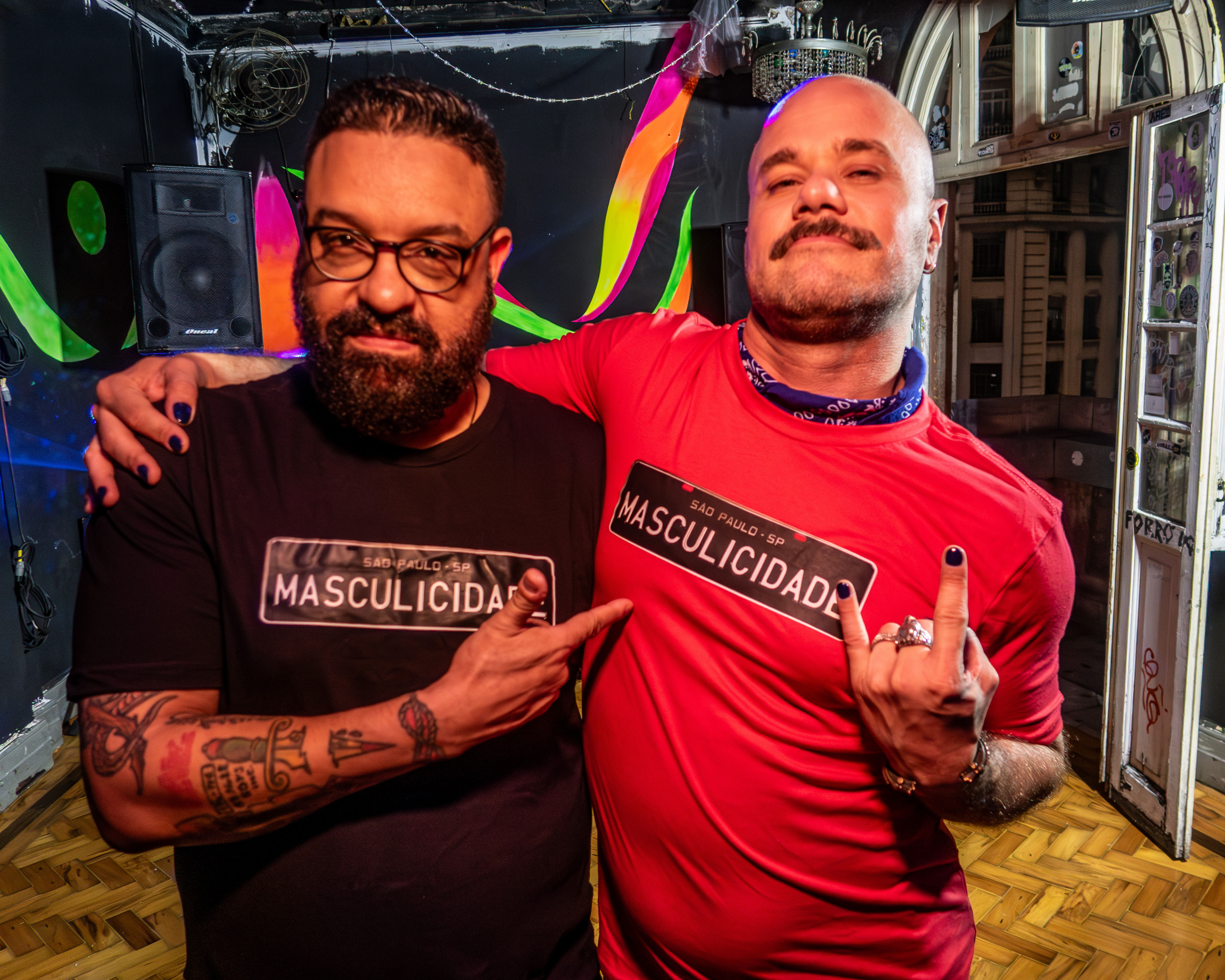 Homem usando camiseta preta Masculicidade abraçando outro homem usando camiseta vermelha Masculicidade.