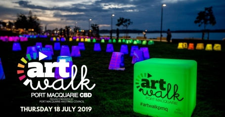 Colorful light cubes on a grassy area at dusk, with text "art walk, Port Macquarie CBD, Thursday 18 July 2019". People in the background.