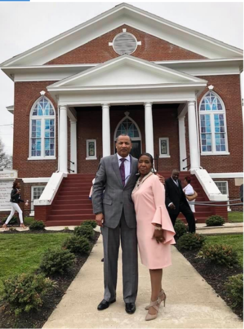 Pastor Barry L. Winston, wearing a dark grey suit, and Dollnita M. Winston, wearing and pastel pink dress stand in the concrete walkway leading to the brick and white trimmed church. The walkway is lined with green bushes and grass.