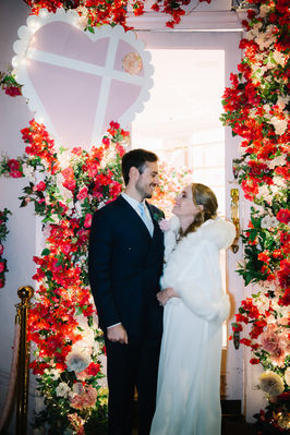 Bride with an elegant updo in a white fur wrap beside her groom under a red rose arch