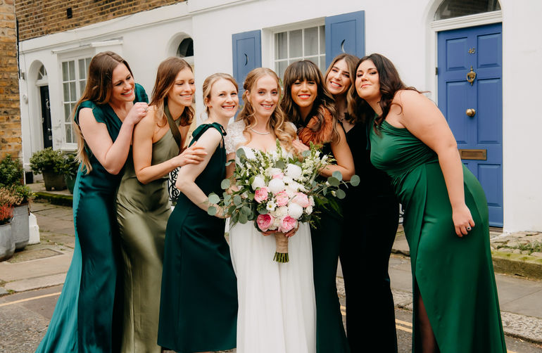 Bride with her six bridesmaids in green dresses, all with soft natural makeup and loose waves, outside a white London townhouse with a blue door