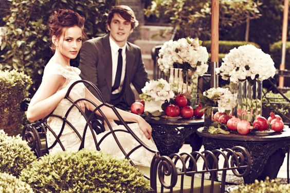 Bride with soft glam makeup and an elegant updo sitting on an iron bench beside her groom in a garden with white flowers and pomegranates