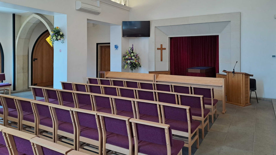 Photo of the interior of Hastings Crematorium Chapel, showing the catafalque, comfy seating, a lectern, a large TV screen, and an old stone Gothic archway.