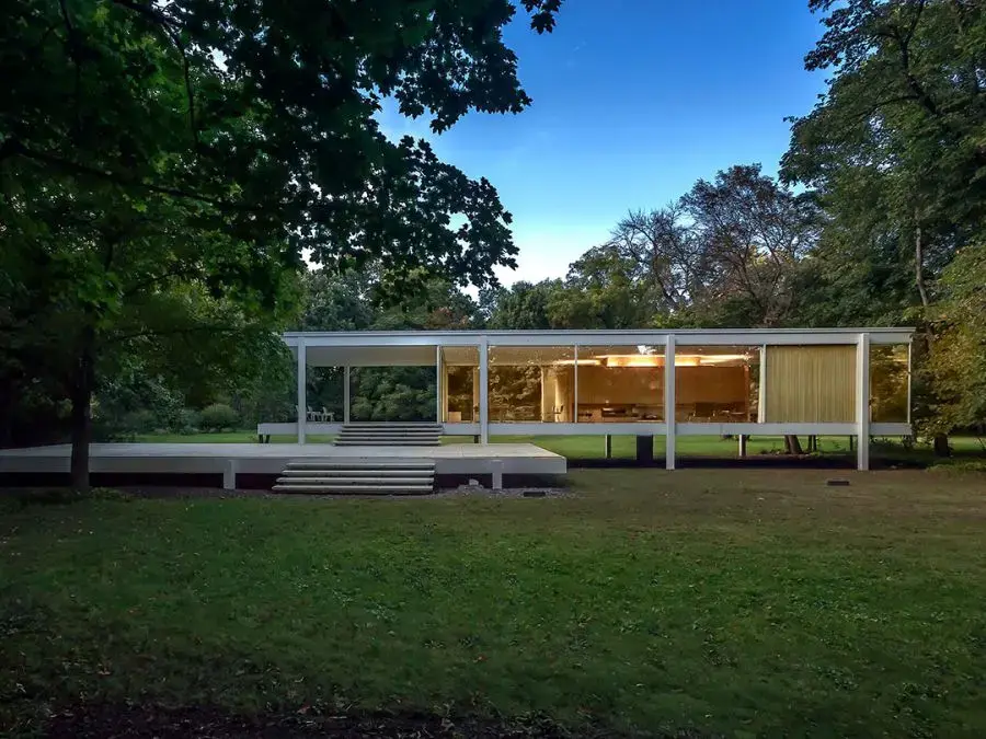 Farnsworth House surrounded by trees, featuring a minimalist white steel frame and large glass walls elevated above the ground.