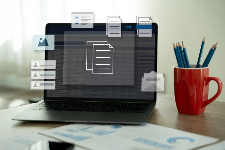 Laptop su una scrivania con un foglio di calcolo e icone di documenti, immagini ed email accanto a una tazza rossa con matite