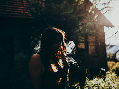 A woman smoking outside near a building with the sun partly shining on her.