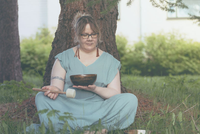 A woman with a singing bowl in her lap sitting at the foot of a tree on the grass