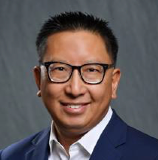 A man wearing a blue jacket and white shirt wearing glasses is posing against a grey background for a headshot