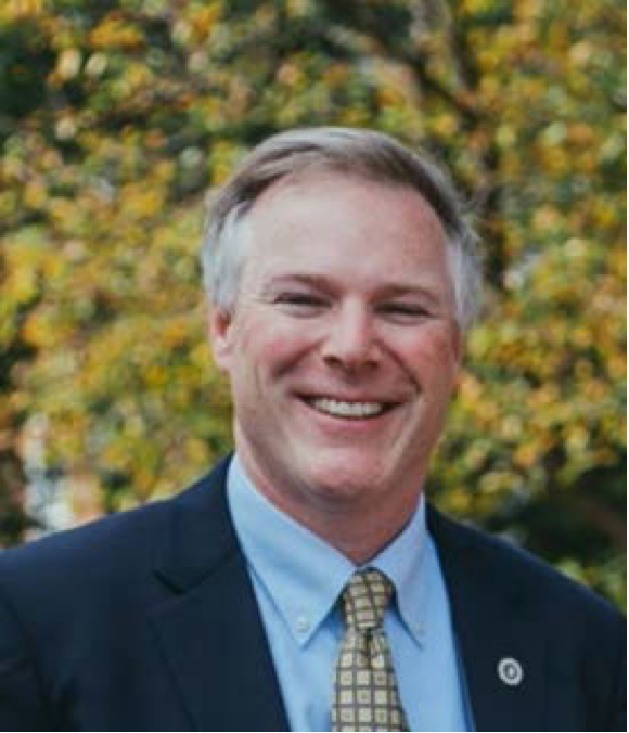 A man in a blue suit and yellow checked tie is smiling and standing in front of a tree