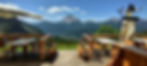 Panoramic terrace of Rifugio Scotter overlooking Mount Pelmo and the Dolomites, with wooden tables, flowers, and clear blue sky.
