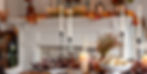 Candles and a vase with autumn leaves decorate a dining table with plates and bread. A fireplace adorned with fall decor is in the background.