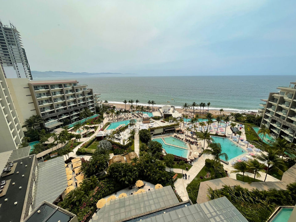 Stunning view from an oceanfront room at Dreams Vallarta Bay, overlooking the resort's vibrant, inviting pools which lead directly to the expansive ocean. The crystal-clear pools are surrounded by lush tropical foliage and plentiful lounge areas, with the calm blue sea extending to the horizon under a bright, clear sky.