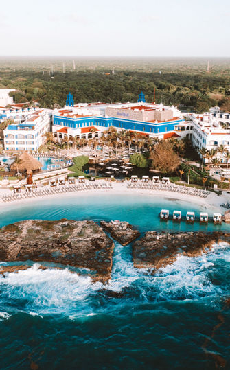 View from the ocean of Hard Rock Riviera Maya Resort, featuring its distinct architecture and plush accommodations. The resort is nestled against a backdrop of lush tropical greenery with two inviting cove beaches framing the scene. Gentle waves lap at the sandy shores under a bright, clear sky, highlighting the resort's tranquil and luxurious setting.