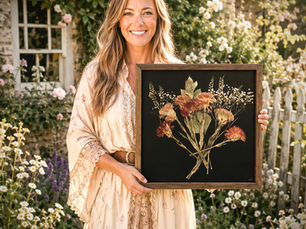 Smiling woman in a garden holding a framed pressed flower bouquet on a black background