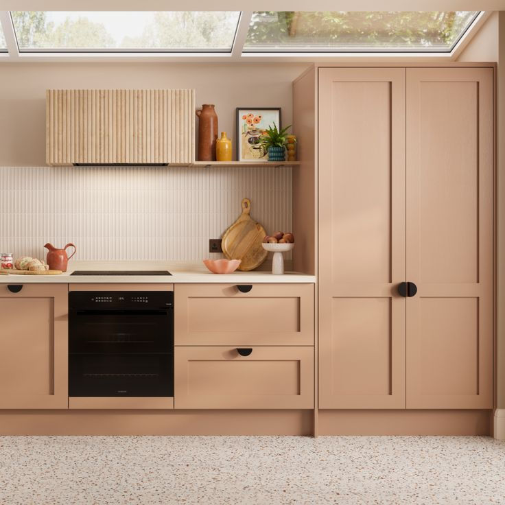Modern kitchen with rose beige cabinets, a black oven, and skylight. Wooden shelves hold decor. Terrazzo floor adds texture and warmth 2026 kitchen trends