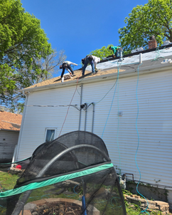 Three workers in safety harnesses are laying new shingles on a steep residential roof. Roofing tools and material are scattered across the surface. Bright blue sky and tree foliage surround the scene, emphasizing the active roof renovation in progress.