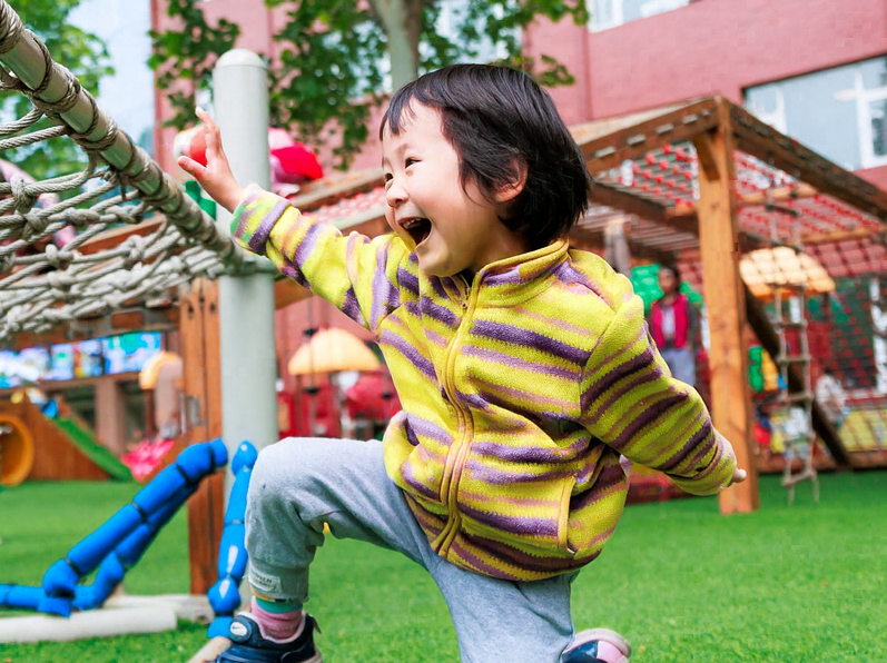 A young child joyfully climbs a rope on a colorful playground, surrounded by swings and slides under a bright blue sky.