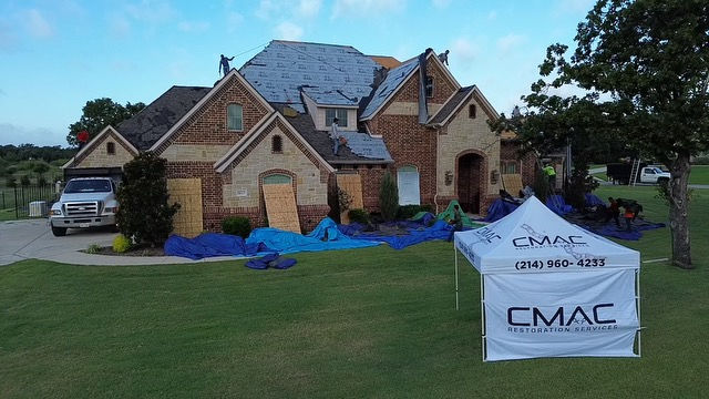Workers are repairing the roof of a brick house. Blue tarps cover the ground. A white tent shows "CMAC Restoration Services" text.