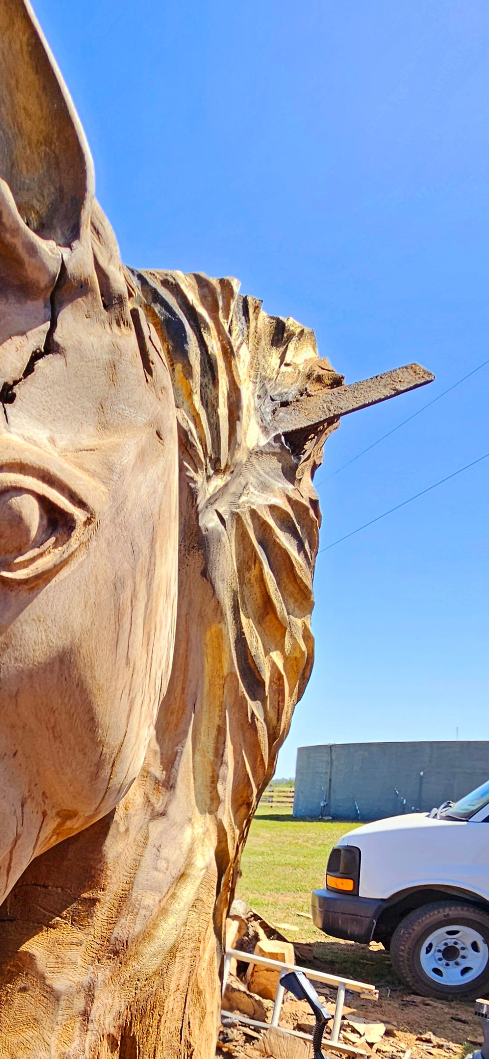 Close-up of a chainsaw horse carving with a horse rasp sticking out of its head against a blue sky. DustyGirl Carver white truck and a building are in the background on grassy land.