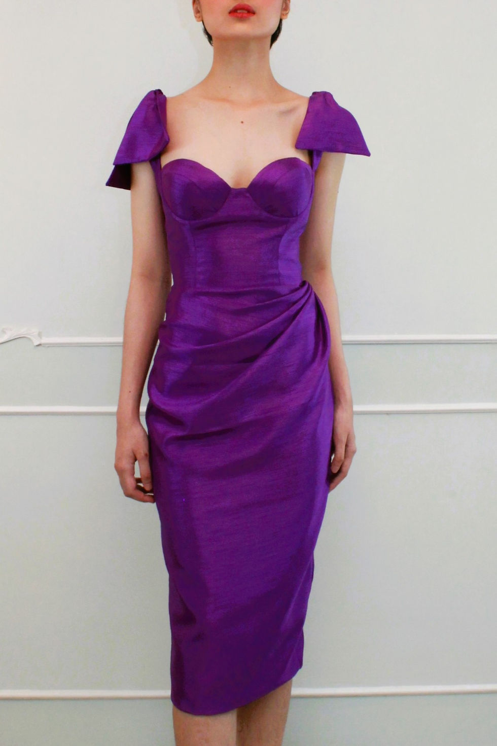 A lady wearing a JACQ purple corset knee length cocktail dress, with bow shoulder and draping on the side of the skirt.