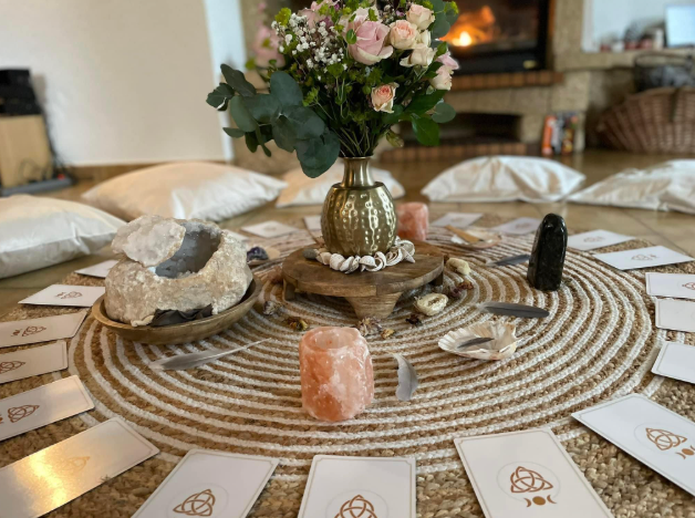 Cartes de tarot en cercle autour de fleurs roses, cristaux et coquillages sur un tapis tressé, dans un salon chaleureux.