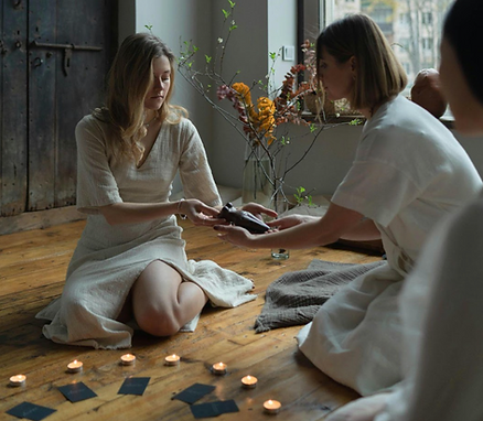 Deux femmes en robes claires s’échangent un petit objet à genoux, dans une pièce chaleureuse avec bougies et cartes au sol.