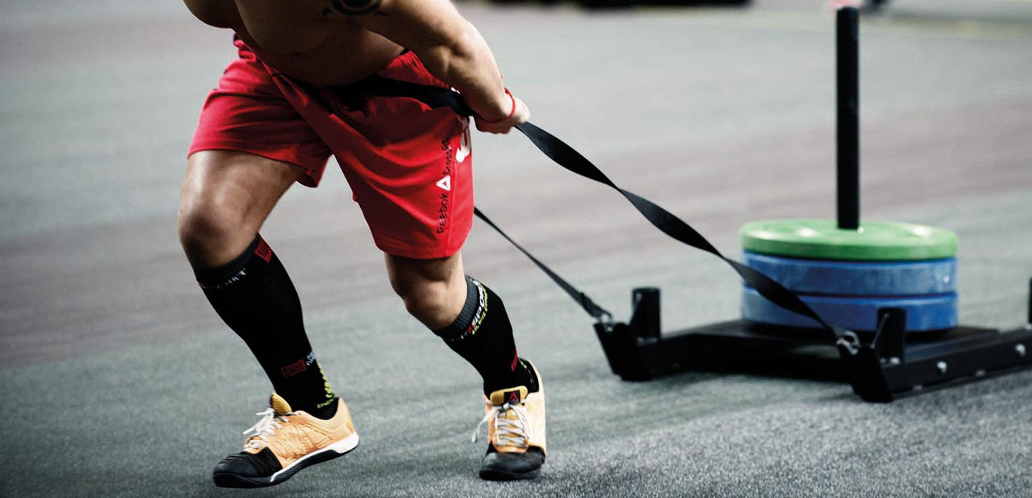 mage montrant un athlète tatoué tirant un traîneau de poids lourd lors d'une compétition sur un revêtement de sol Caoutchouc SPORTEC de finition Gris clair fourni par Sport Surfaces à Casablanca. Idéal pour entraînement fonctionnel et Box de Crossfit, optimisant la résistance à l'abrasion du traîneau et la stabilité pour l'effort.