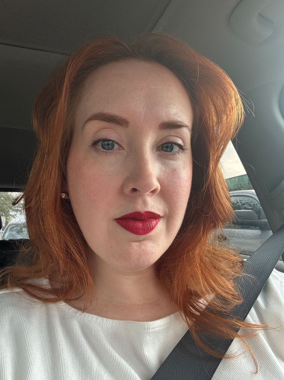 White woman with blue eyes and wavy red hair is sitting casually in a vehicle. She wears red lipstick and a white shirt.