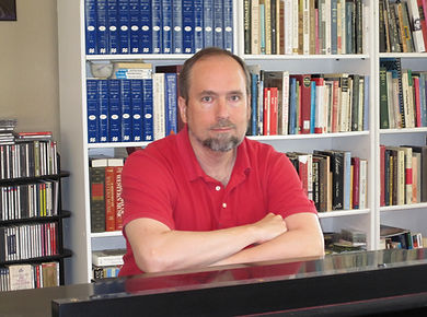 Dr. Stuart Foster seated at the piano in his studio, in front of the New Grove Dictionary and many learned music books.