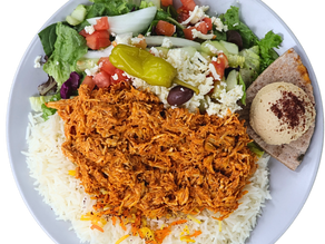 Plate with shredded chicken over rice, Greek salad with olives, pepper, feta, pita bread, and hummus. Vibrant, fresh meal setting.