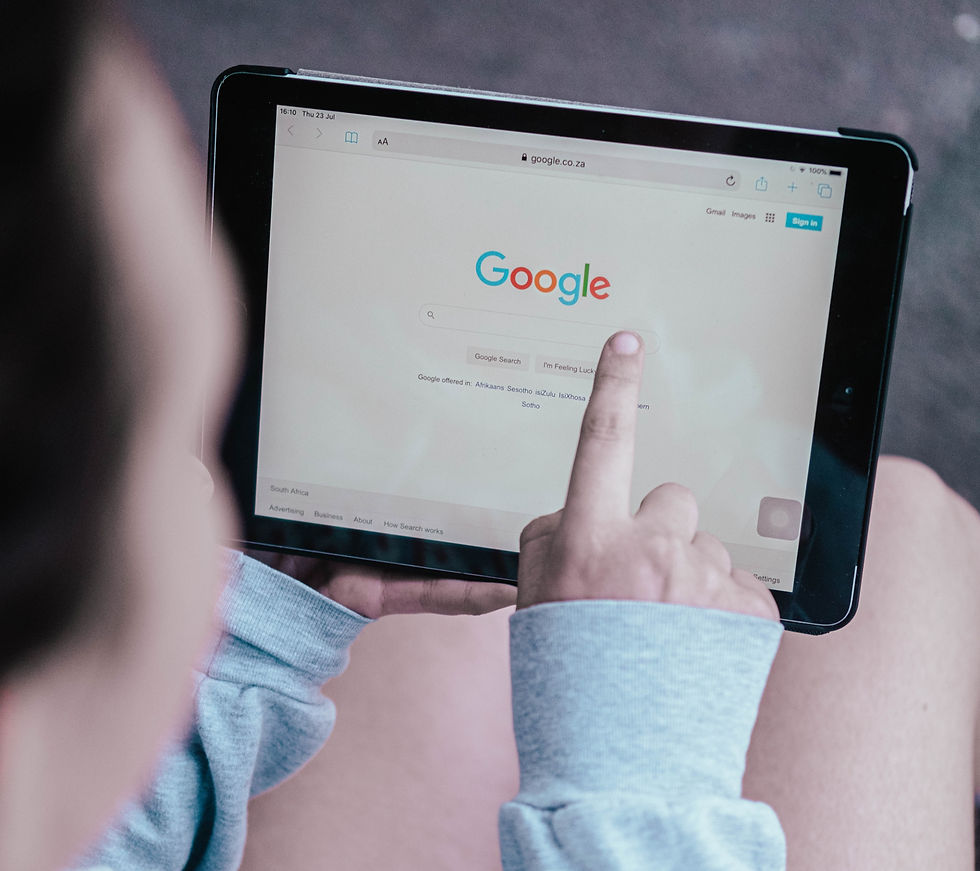 Person in a gray sweater using a tablet with Google homepage on screen. Finger points at the search bar. Casual setting.