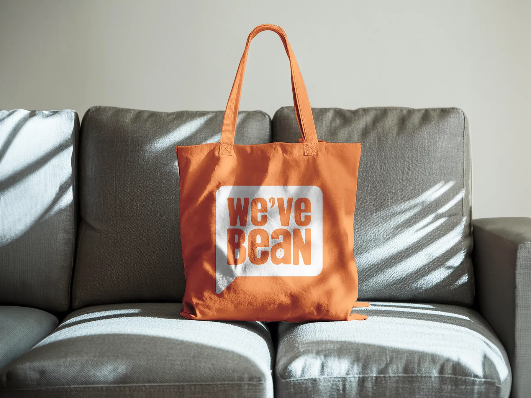 Orange tote bag on a grey couch with the "We've Bean" logo printed in white and orange.