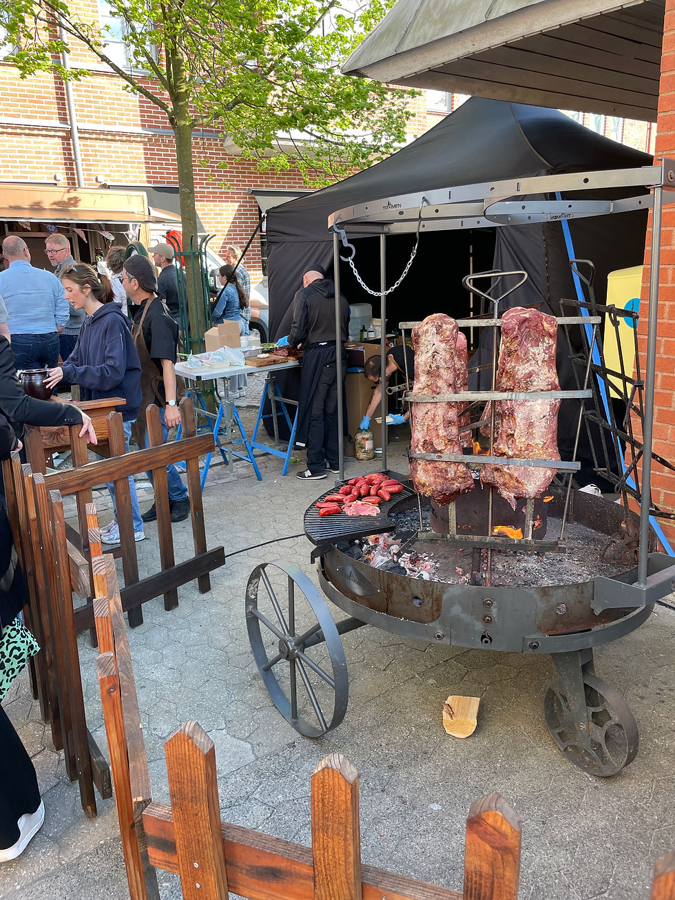 Folk samlas vid ett stånd med en grill, där kött tillagas. Tält och träd i bakgrunden. Solig dag, avslappnad atmosfär.