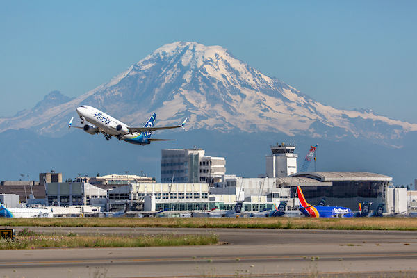 An Alaska Airlines plane taking off in front of Seattle-Tacoma International Airport on a sunny clear day. A snow-covered Mount Rainier is prominent in the background.